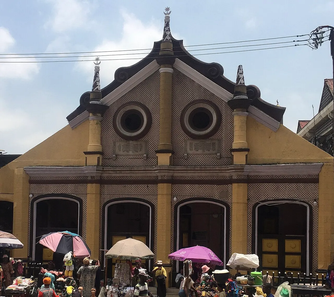 Yellow and white facade of a mosque with a curved roof, crescent moon finials and two round windows, with market stalls and colorful umbrellas in front.