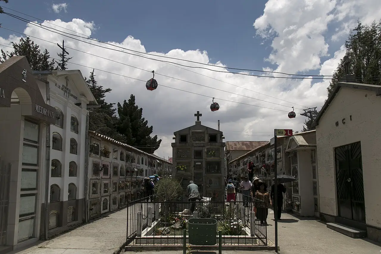 Corredor do Cementerio General, com nichos nas paredes, uma capela com cruz ao fundo e cabos de teleférico no céu.