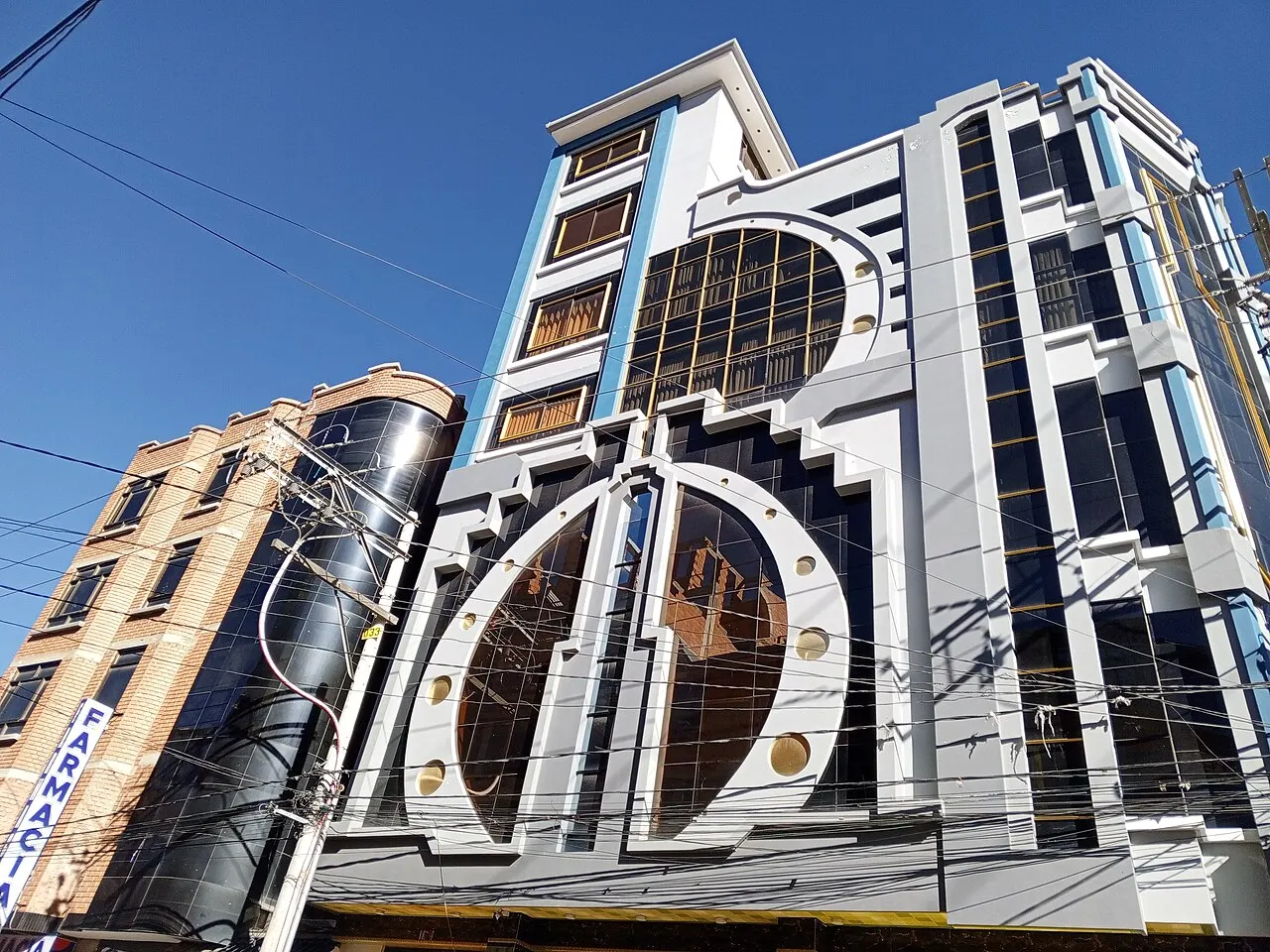 Fachada de um cholet de El Alto vista de baixo: vidro espelhado escuro, molduras brancas em desenho geométrico e discos dourados, com fios elétricos cruzando a frente.
