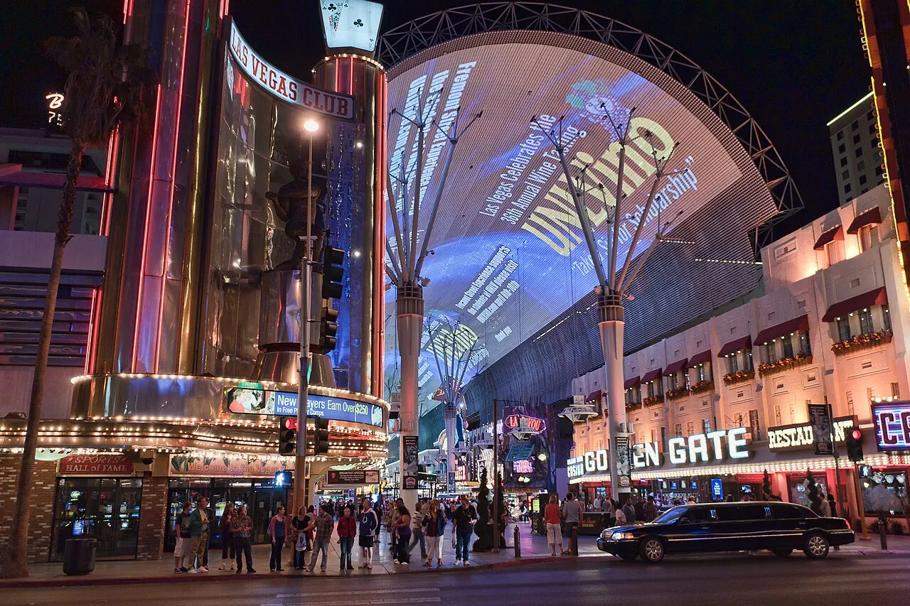 Uma rua coberta por uma grande abóbada de telas de LED exibindo imagens coloridas, ladeada por fachadas de cassino com letreiros de neon como "Las Vegas Club" e "Golden Gate", pessoas caminhando à noite.