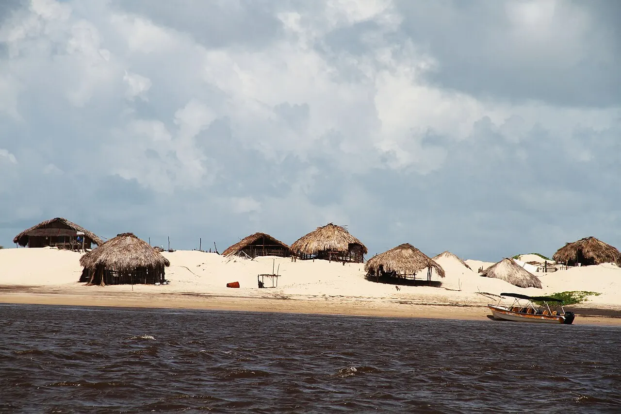 Cabanas com telhado de palha sobre a faixa de areia de Caburé, vistas do rio Preguiças, com um barco na margem e céu nublado.