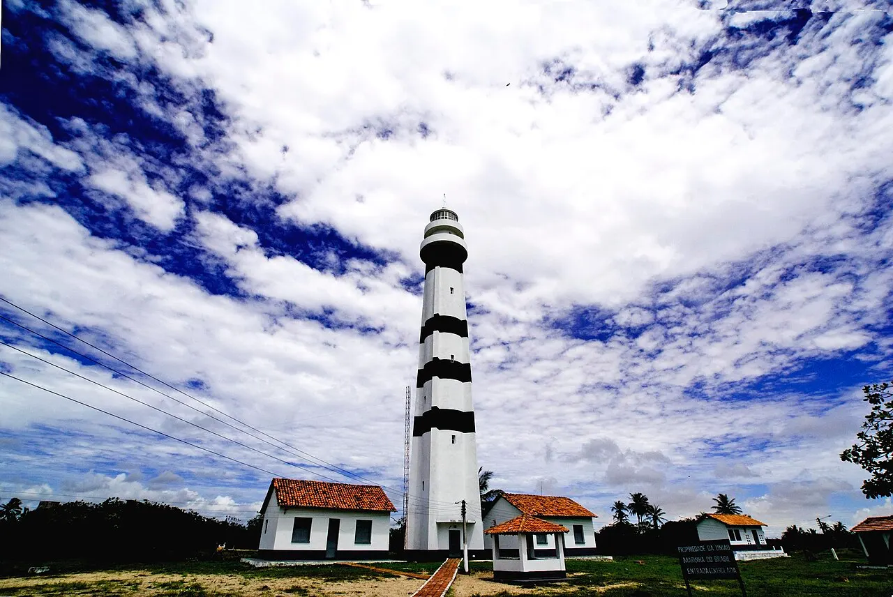 O Farol de Preguiças, uma torre branca com faixas pretas, com casas brancas de telhado vermelho ao pé, sob céu azul com muitas nuvens.
