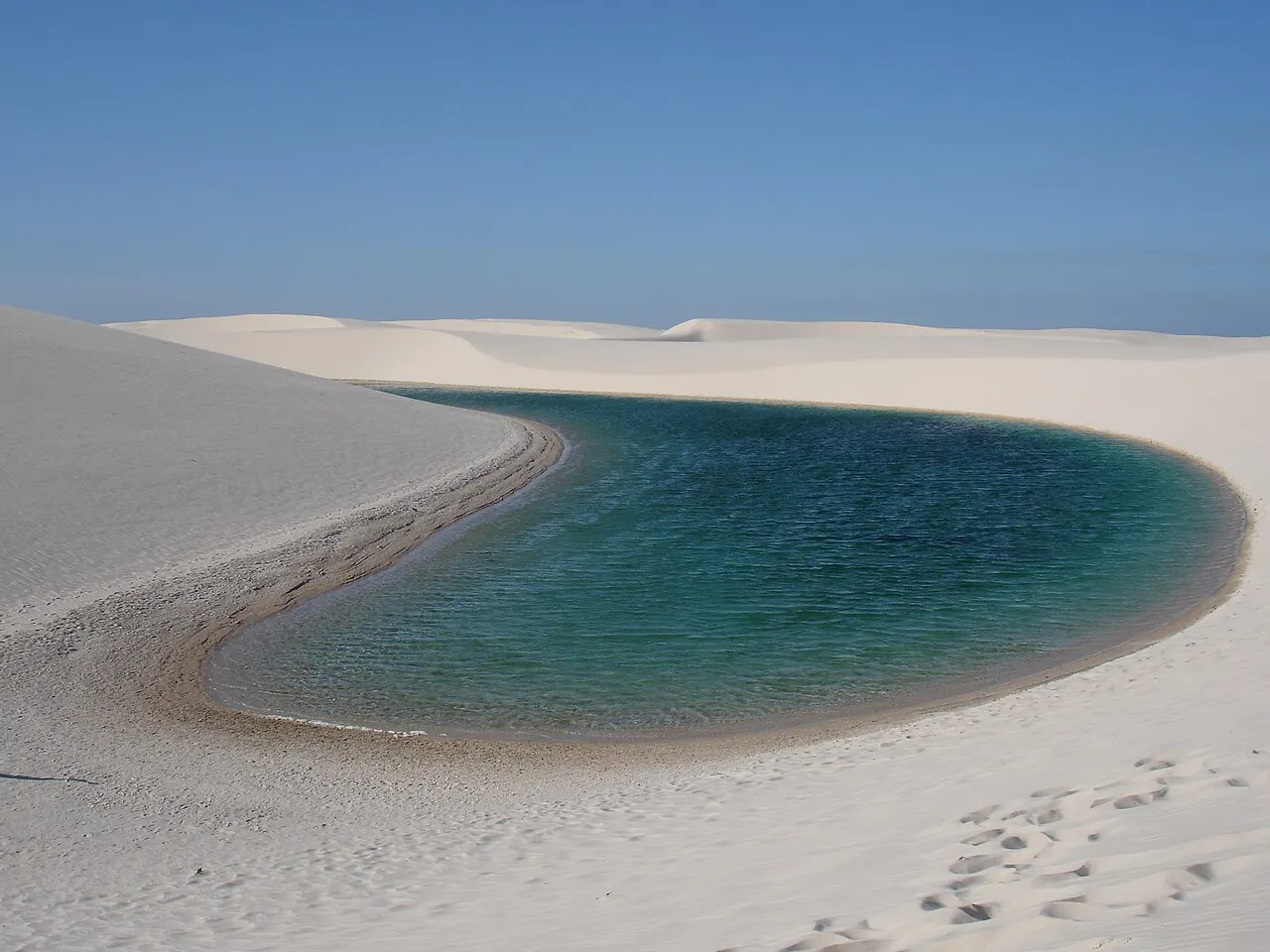 Uma lagoa de água azul-esverdeada encaixada entre dunas de areia branca, com pegadas na areia em primeiro plano e céu azul.