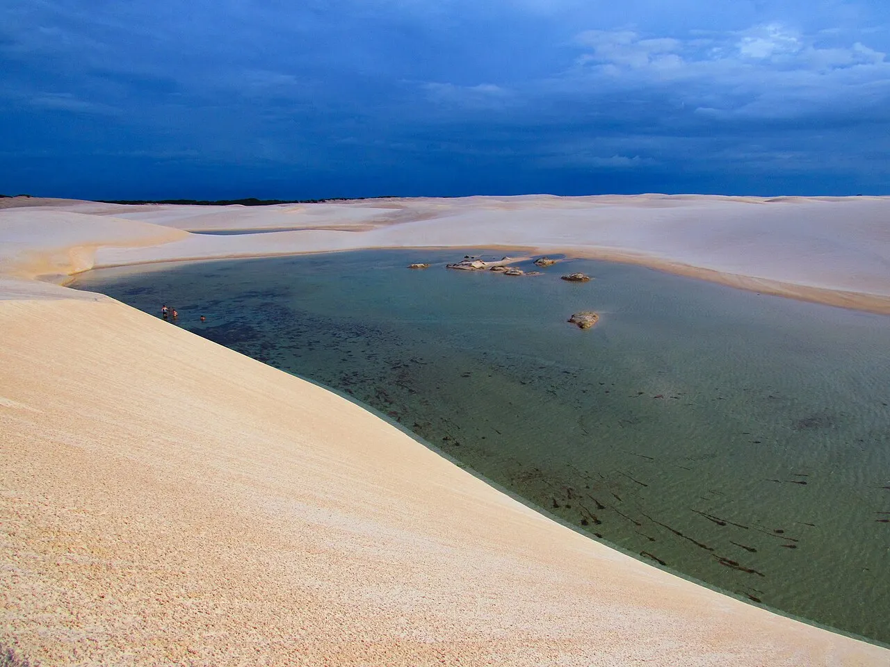 A Lagoa Bonita, uma lagoa de água escura e transparente entre dunas brancas, sob um céu azul carregado de nuvens.