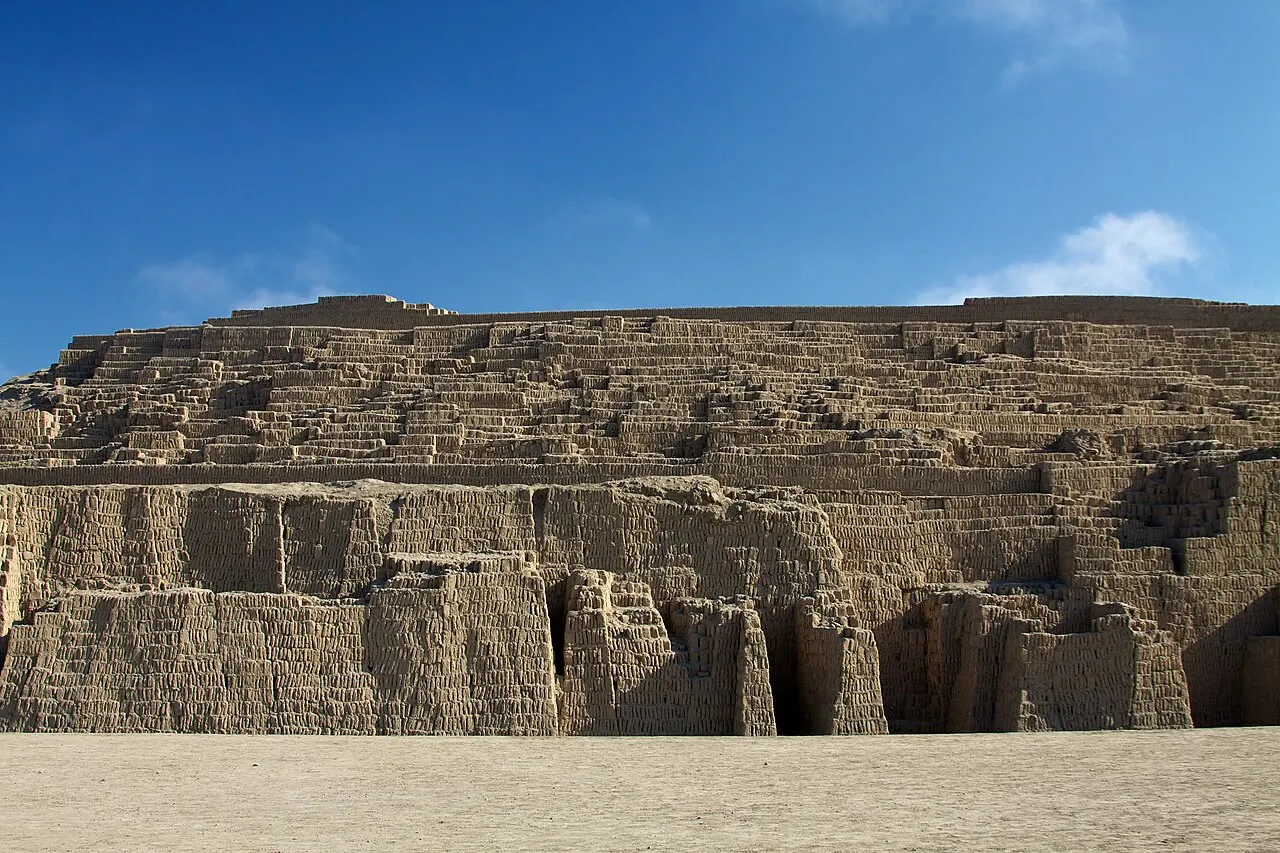 Pirâmide escalonada de tijolos de adobe postos de pé, em degraus largos, sob céu azul, com o pátio de areia em primeiro plano.