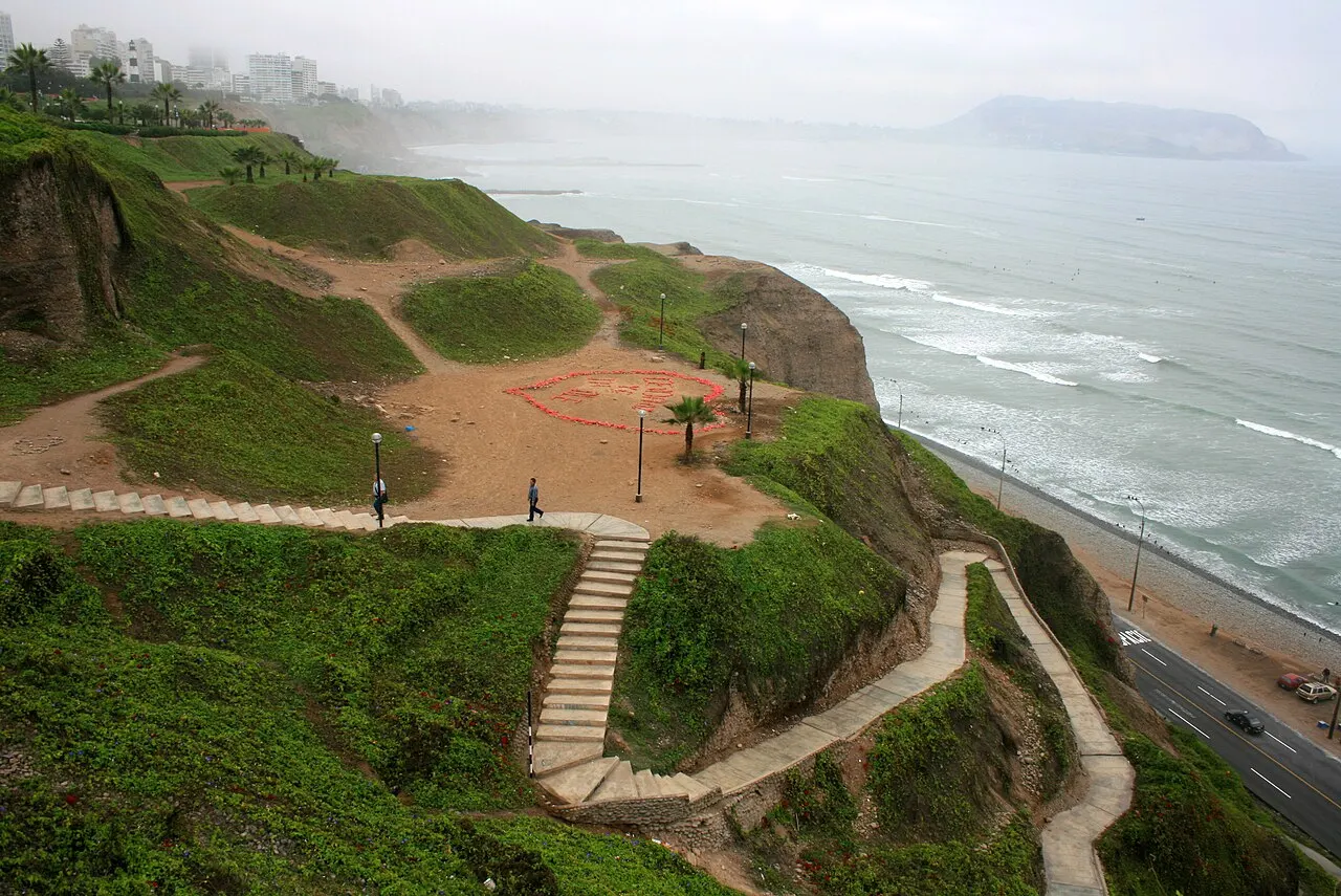 Um penhasco gramado à beira-mar, com escadas e caminhos sinuosos descendo até a orla, um desenho de coração feito de flores no chão, prédios ao longe encobertos por neblina e ondas quebrando na praia abaixo.