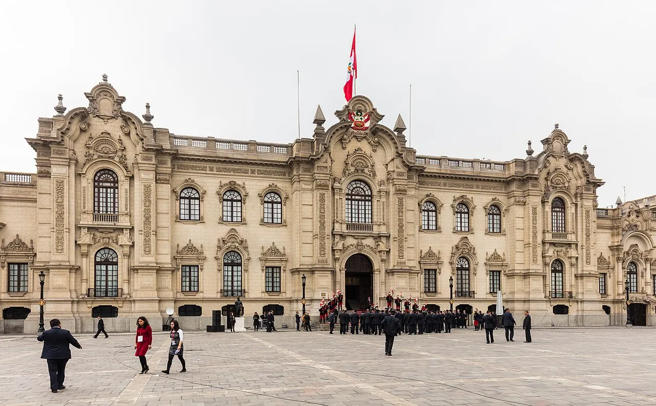 Fachada do Palacio de Gobierno na praça, com a bandeira do Peru e pessoas no piso de pedra.