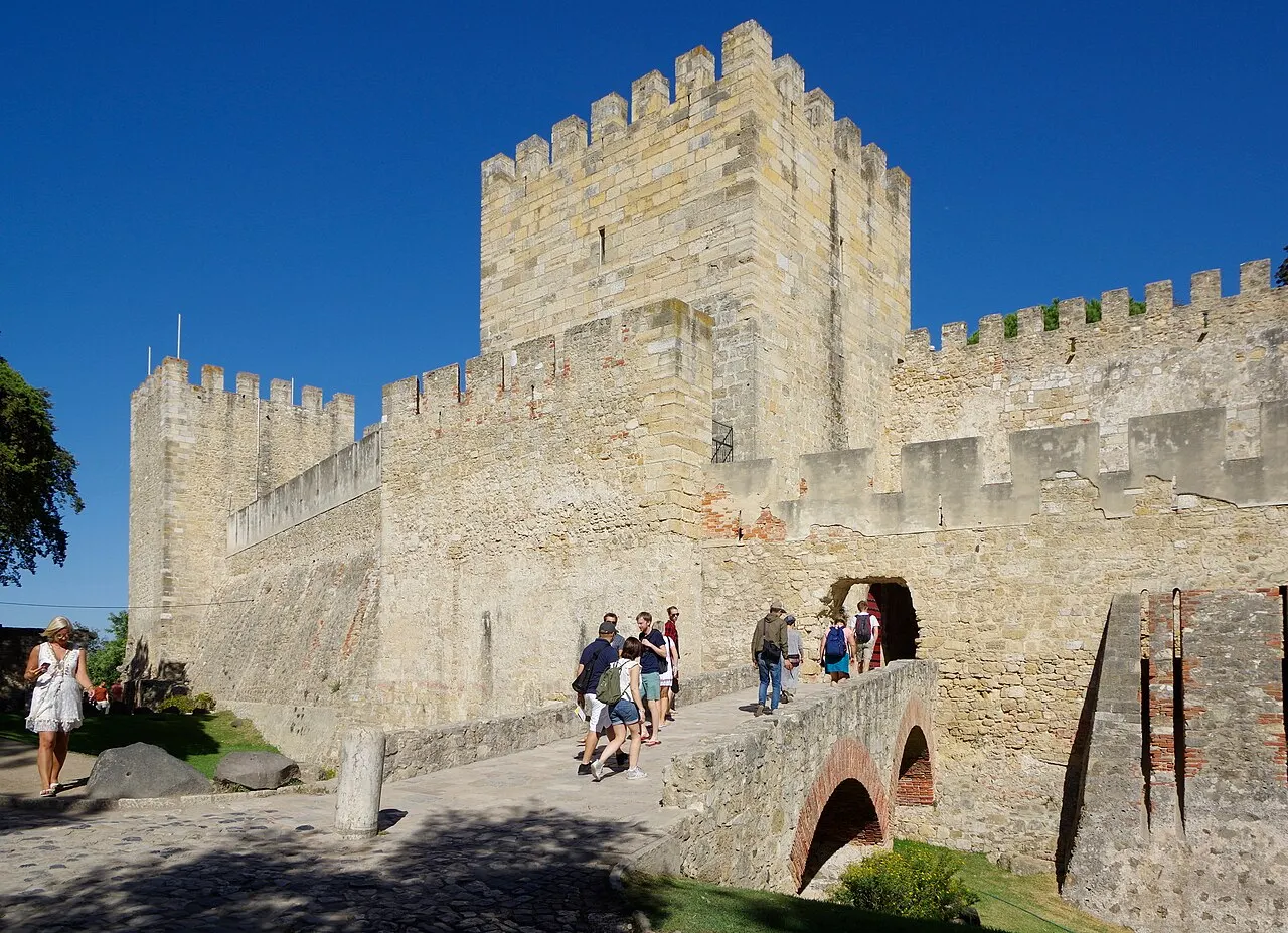 Uma torre de pedra ameada com muralhas ao redor, um fosso e uma ponte de arcos de tijolo por onde turistas caminham em direção a uma passagem na muralha, sob céu azul intenso.