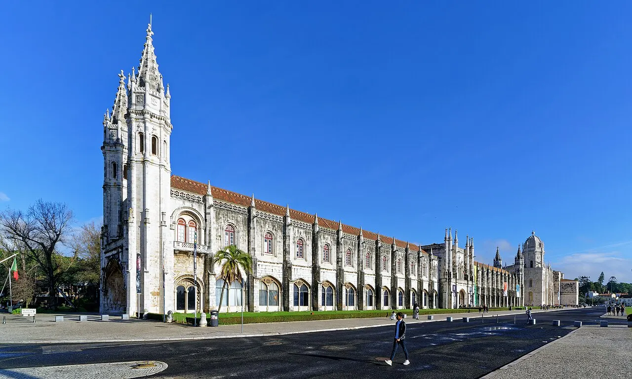 Fachada sul do Mosteiro dos Jerónimos, em Lisboa, de pedra clara, com a torre e o portal manuelino, sob céu azul.