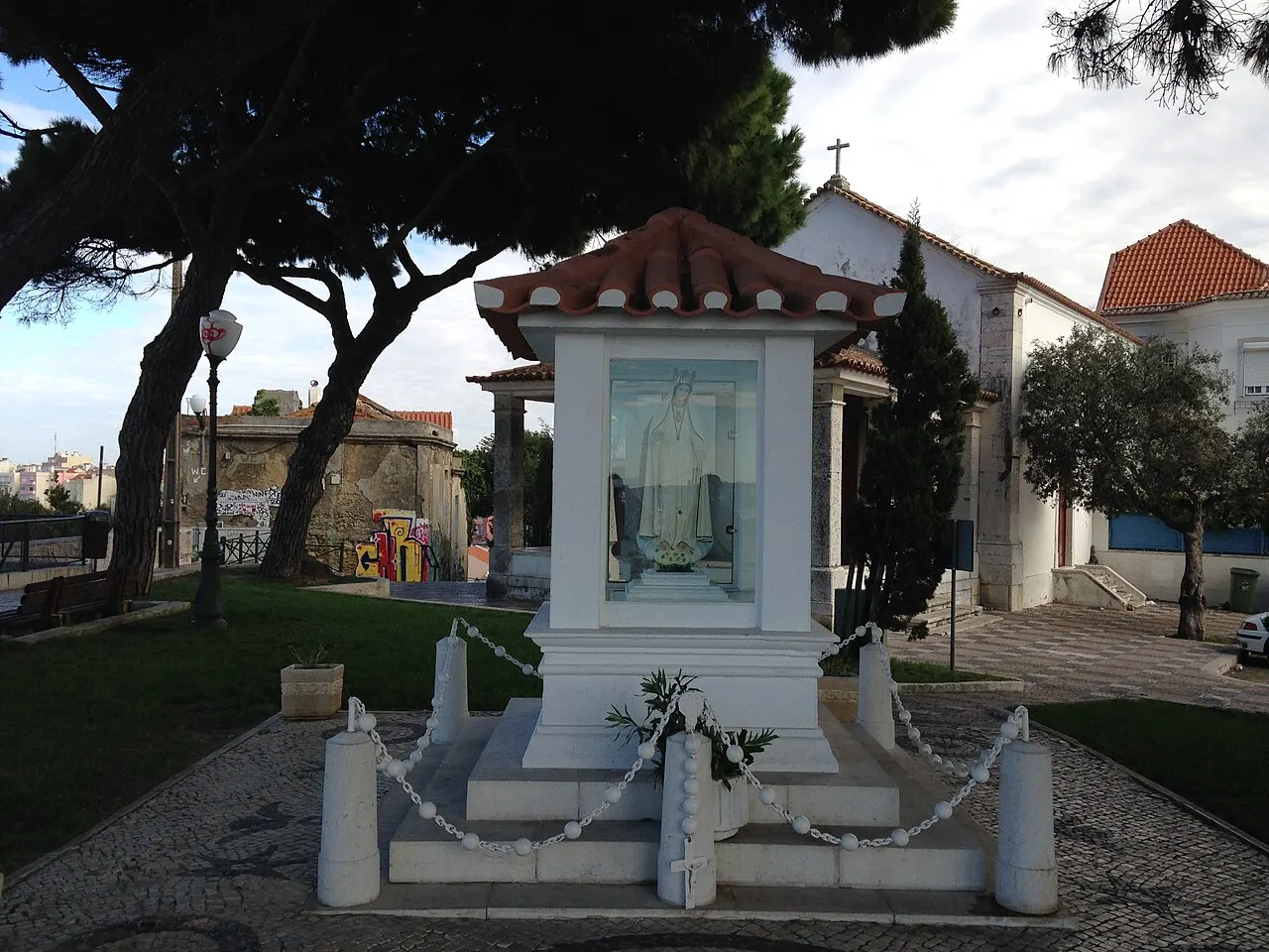 Oratório branco com telhado e painel de azulejo, sob árvores, no Miradouro da Senhora do Monte, em Lisboa.