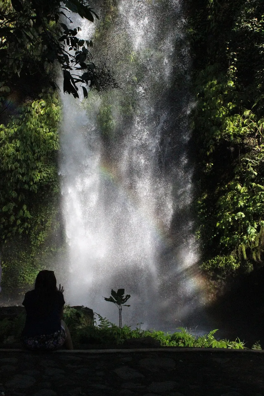 La cascade de Sendang Gile tombant d'une paroi couverte de végétation, avec de la bruine et un léger arc-en-ciel, et la silhouette d'une personne de dos au belvédère.