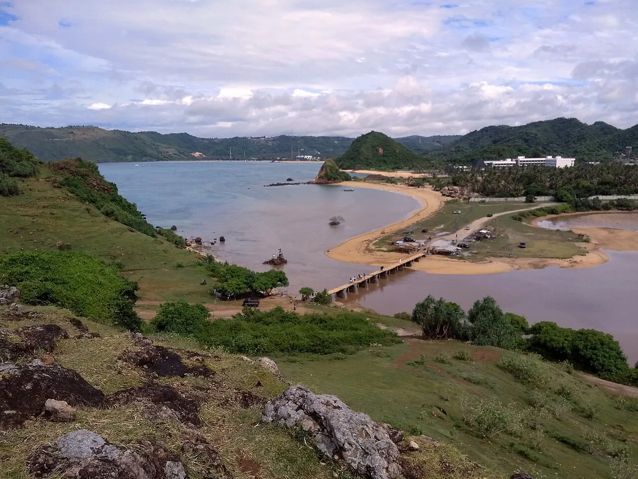 Pente d'herbe et de rochers au premier plan et, en contrebas, une baie peu profonde avec une langue de sable, un petit pont, des collines vertes et des bâtiments au loin.