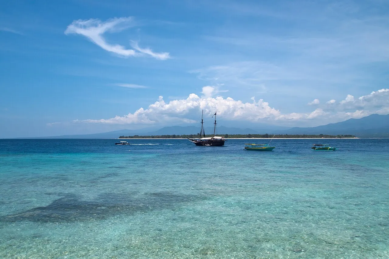 Mer transparente et turquoise au-dessus des récifs, avec un voilier à deux mâts et de petits bateaux au mouillage devant la bande basse et verte de Gili Air, et les montagnes de Lombok derrière.