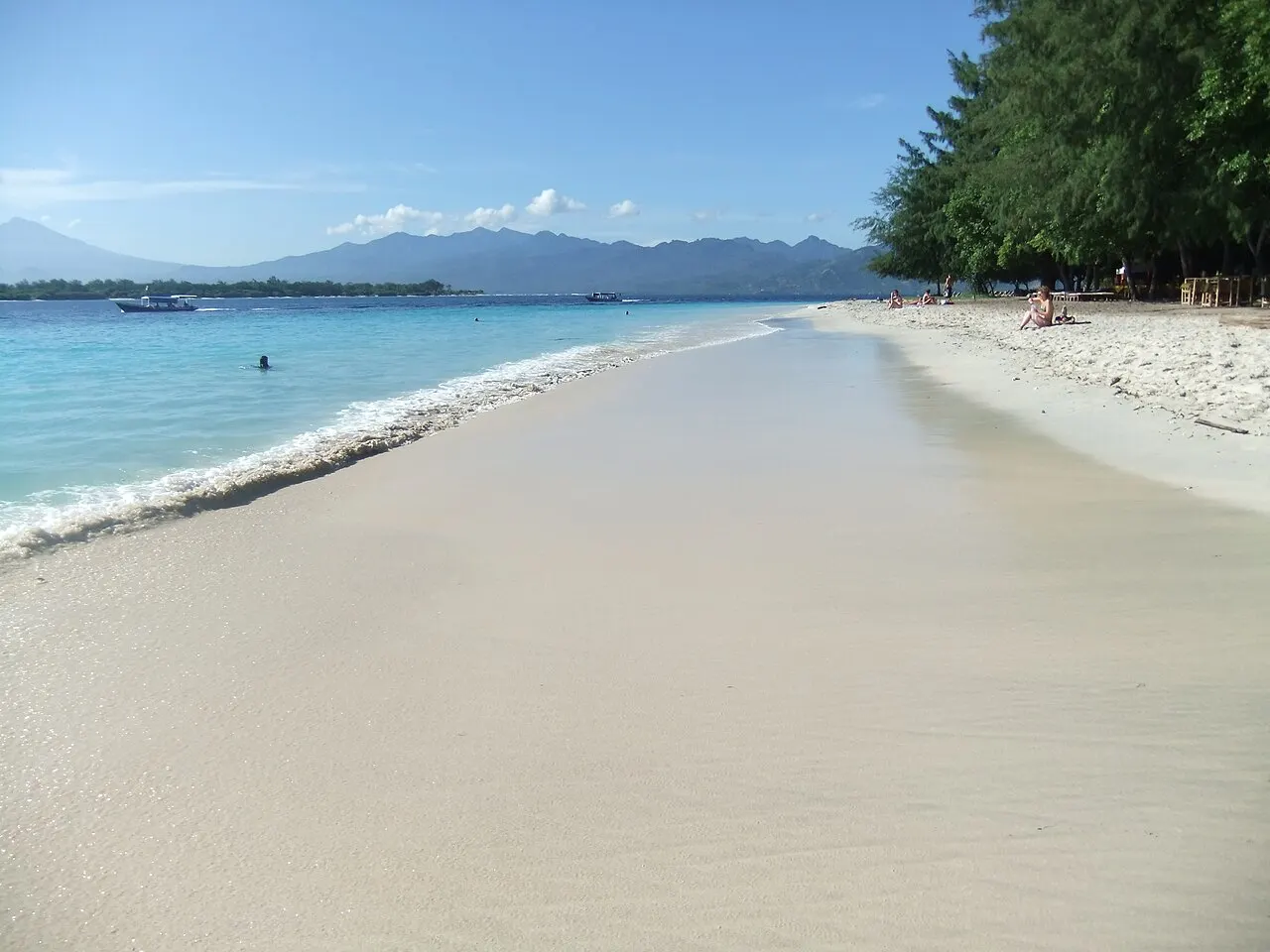 Une large bande de sable blanc à Gili Trawangan, mer peu profonde bleu clair à gauche, arbres à droite et les montagnes de Lombok à l'horizon.