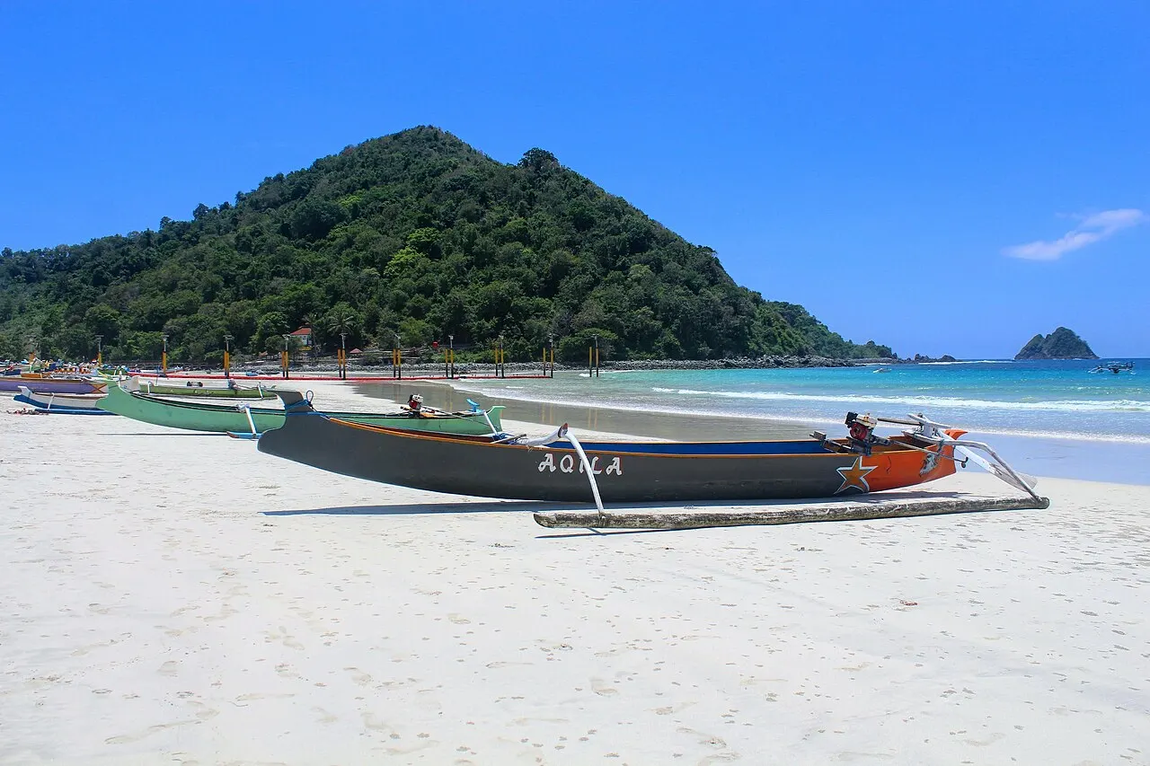 Barques de pêche en bois à balanciers posées sur le sable blanc de Selong Belanak, la mer bleue et une colline boisée de l'autre côté de la baie.