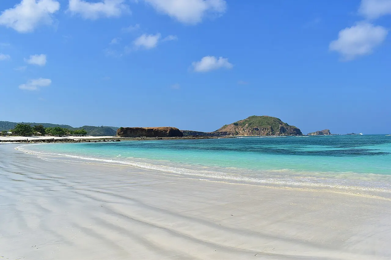 Le sable clair de Tanjung Aan strié par les vagues, la mer turquoise et, au fond, un rocher bas et une colline verte au bout de la baie, sous un ciel bleu nuageux.