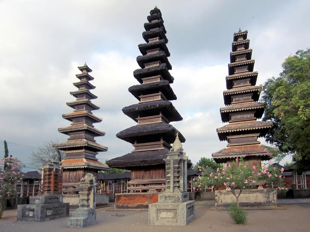 Trois tours meru en bois aux nombreux toits sombres superposés dans la cour en terre du Pura Meru, avec de petits sanctuaires en pierre et des arbustes en fleurs.