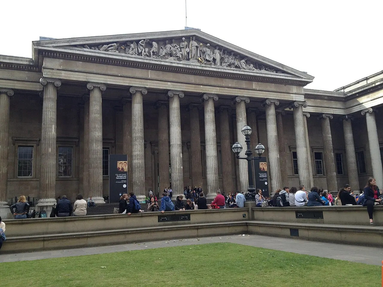 Fachada do British Museum com sua longa colunata jônica e frontão esculpido, vista do pátio gramado, com visitantes sentados nos degraus e no murinho.