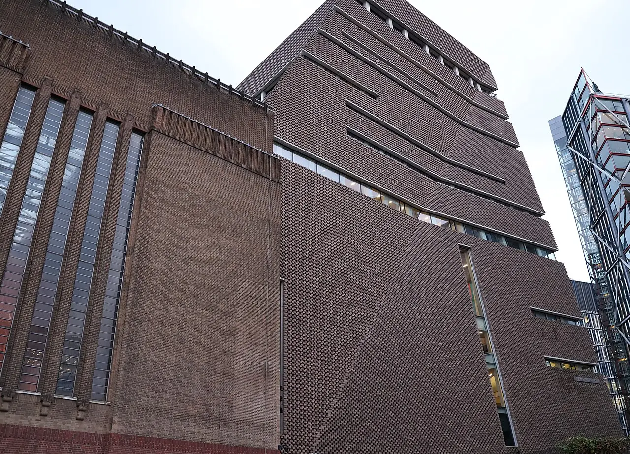 Fachada angular de tijolos da Tate Modern, com faixas de janelas e volumes escalonados, ao lado de um edifício de vidro sob céu claro.