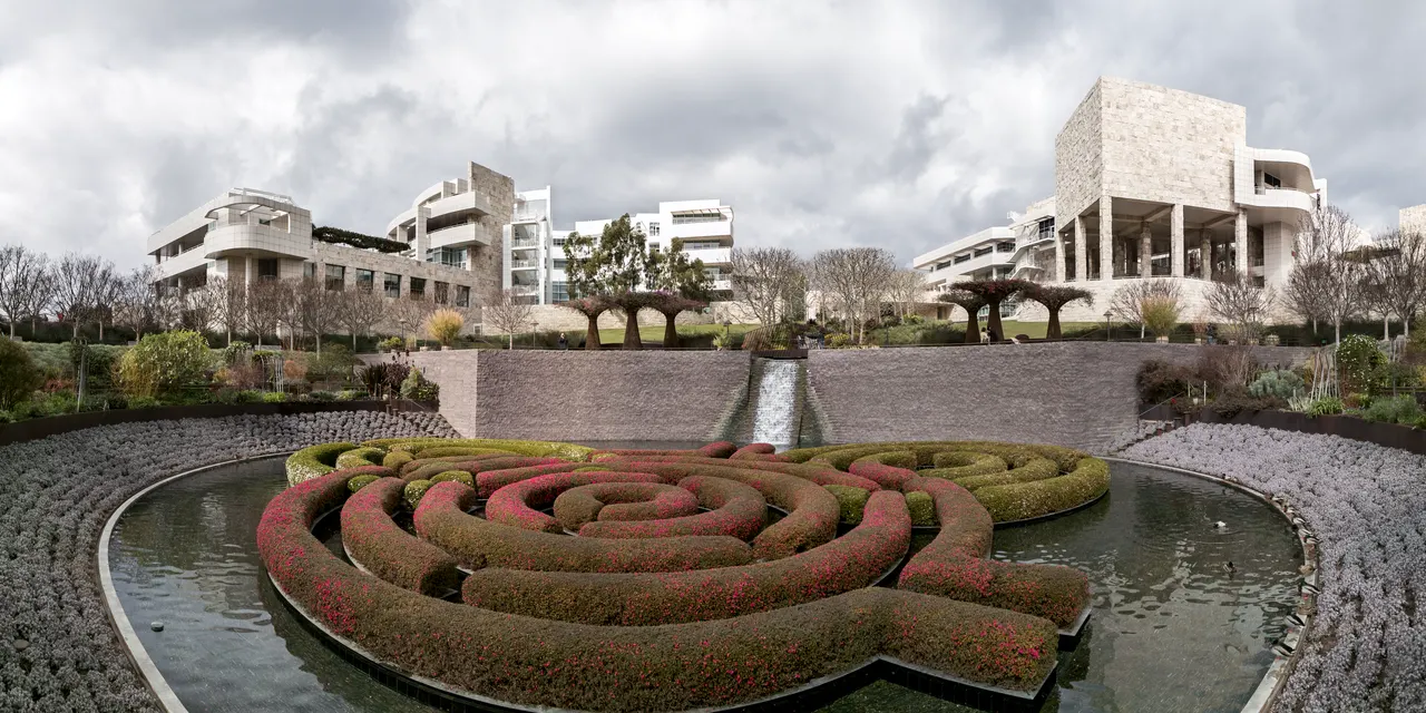Um jardim circular com sebes podadas em espiral e flores vermelhas, cercado por água, com pavilhões de pedra clara de arquitetura moderna ao fundo e árvores pequenas ao redor, sob céu nublado.