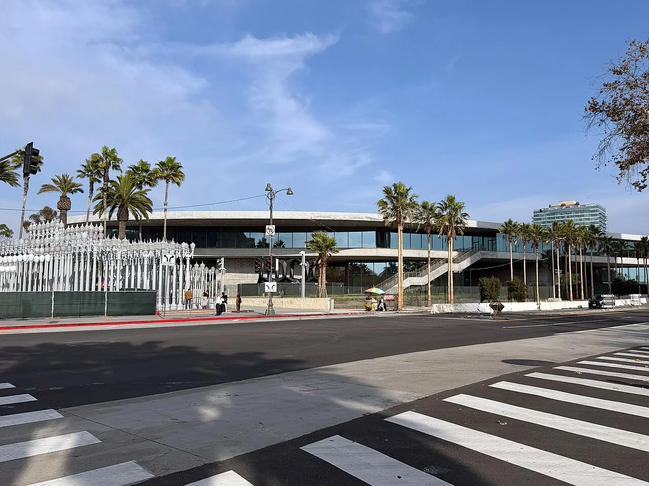 Prédio longo do LACMA com a instalação de postes brancos à esquerda e palmeiras na calçada.