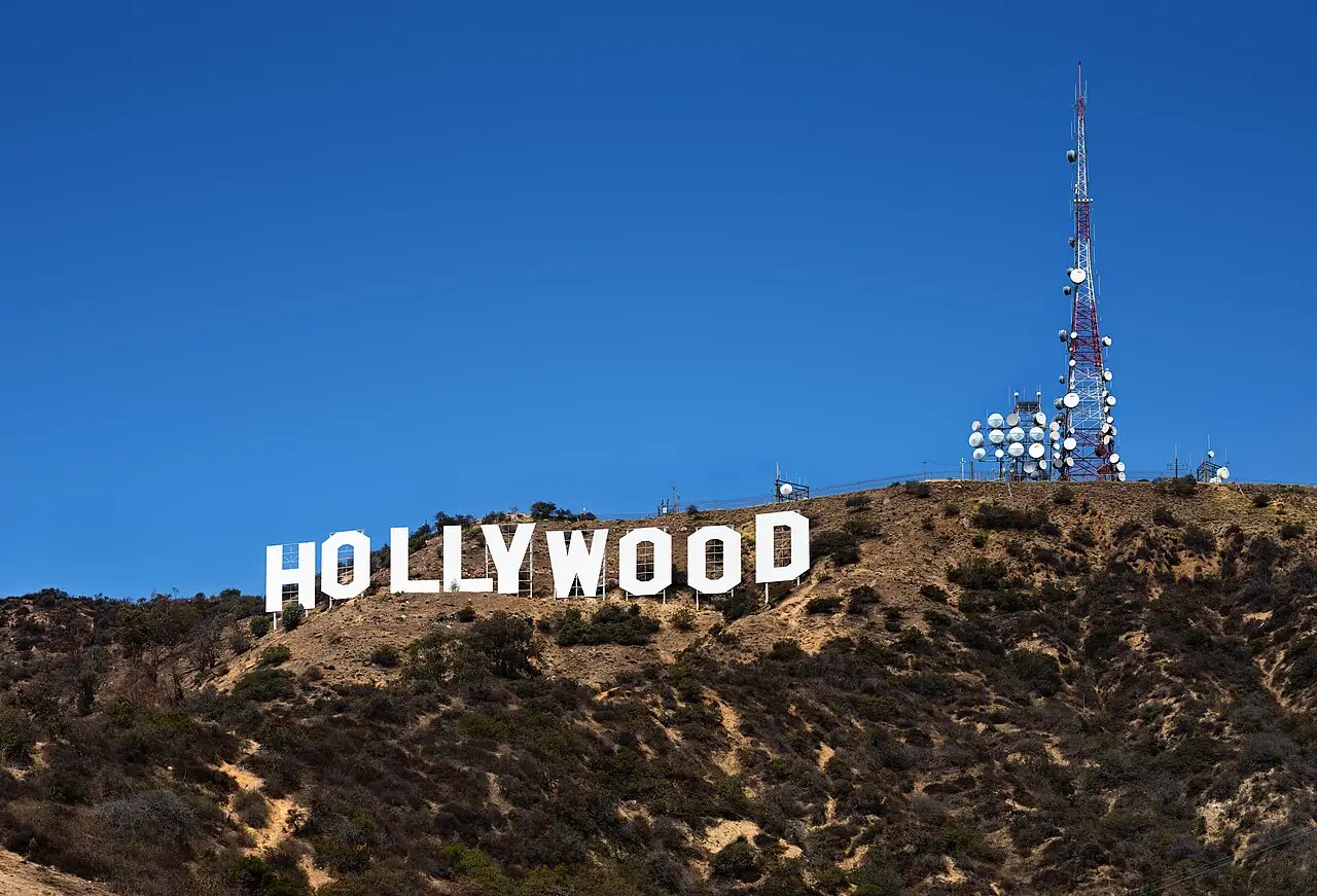 Letras brancas do letreiro de Hollywood na encosta seca, com a torre de rádio no alto do morro e céu azul.