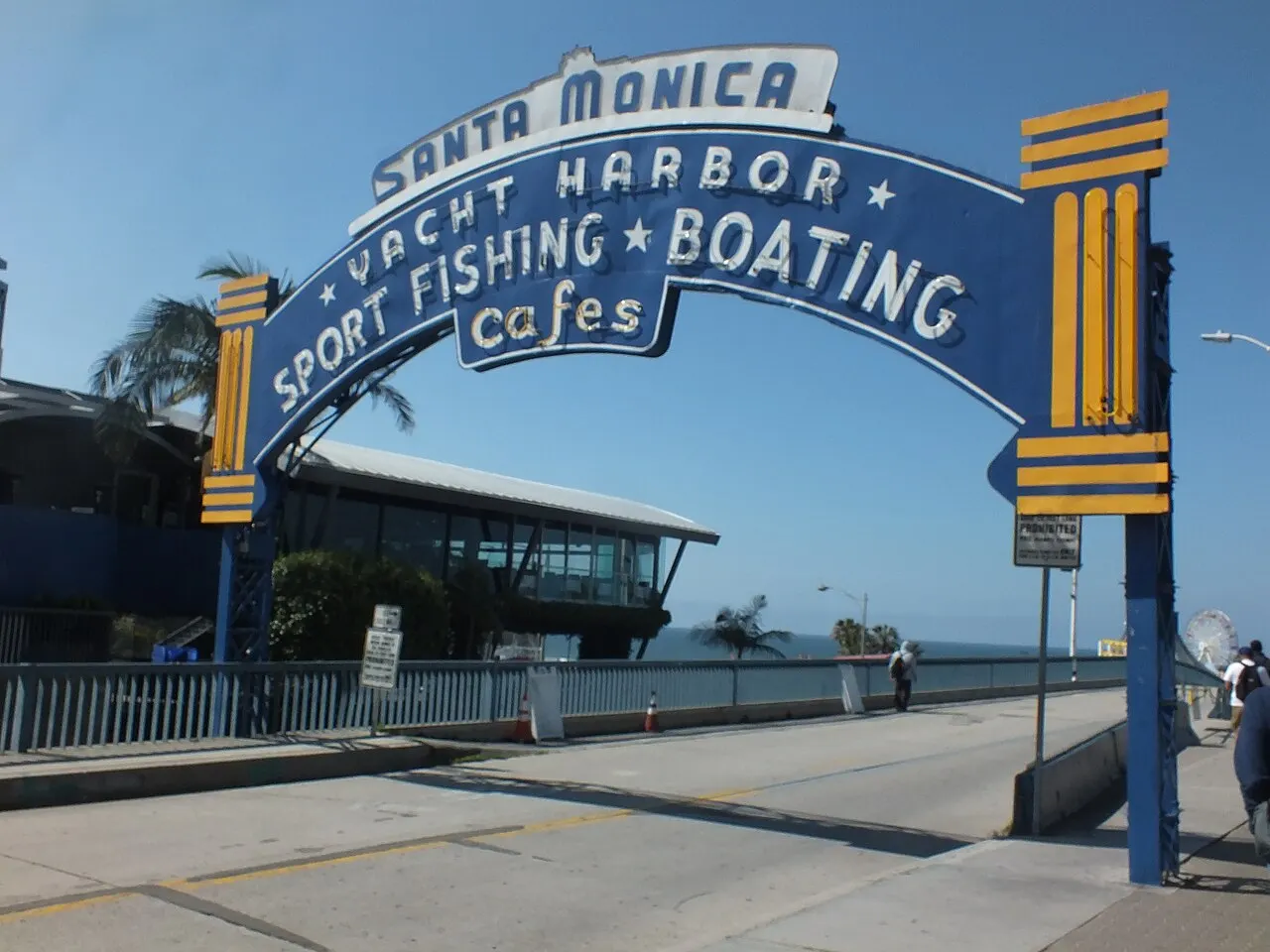 Letreiro em arco da entrada do Píer de Santa Monica, azul e amarelo, sob céu azul.