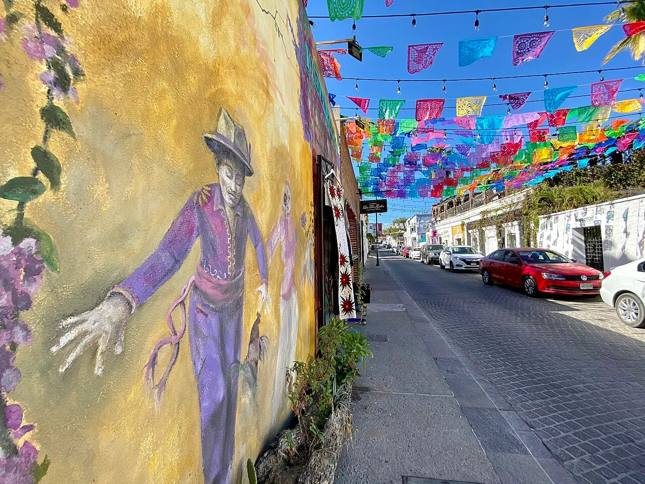 Rua de paralelepípedos com um mural colorido de um homem de chapéu na parede à esquerda, bandeirinhas de papel picado no alto e carros estacionados.