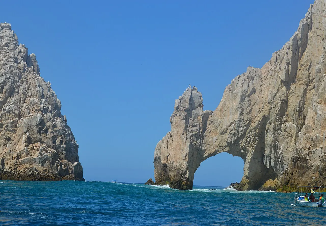 Arco de rocha clara sobre o mar azul, ao lado de um penhasco pontiagudo, com um barco pequeno perto da base e céu limpo.