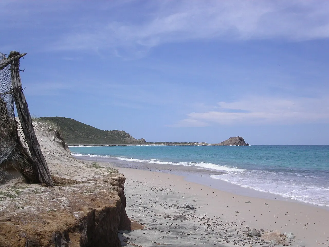 Praia de areia com ondas pequenas e mar azul-claro, um barranco de terra e uma cerca velha à esquerda, e um morro com ilhota ao fundo.