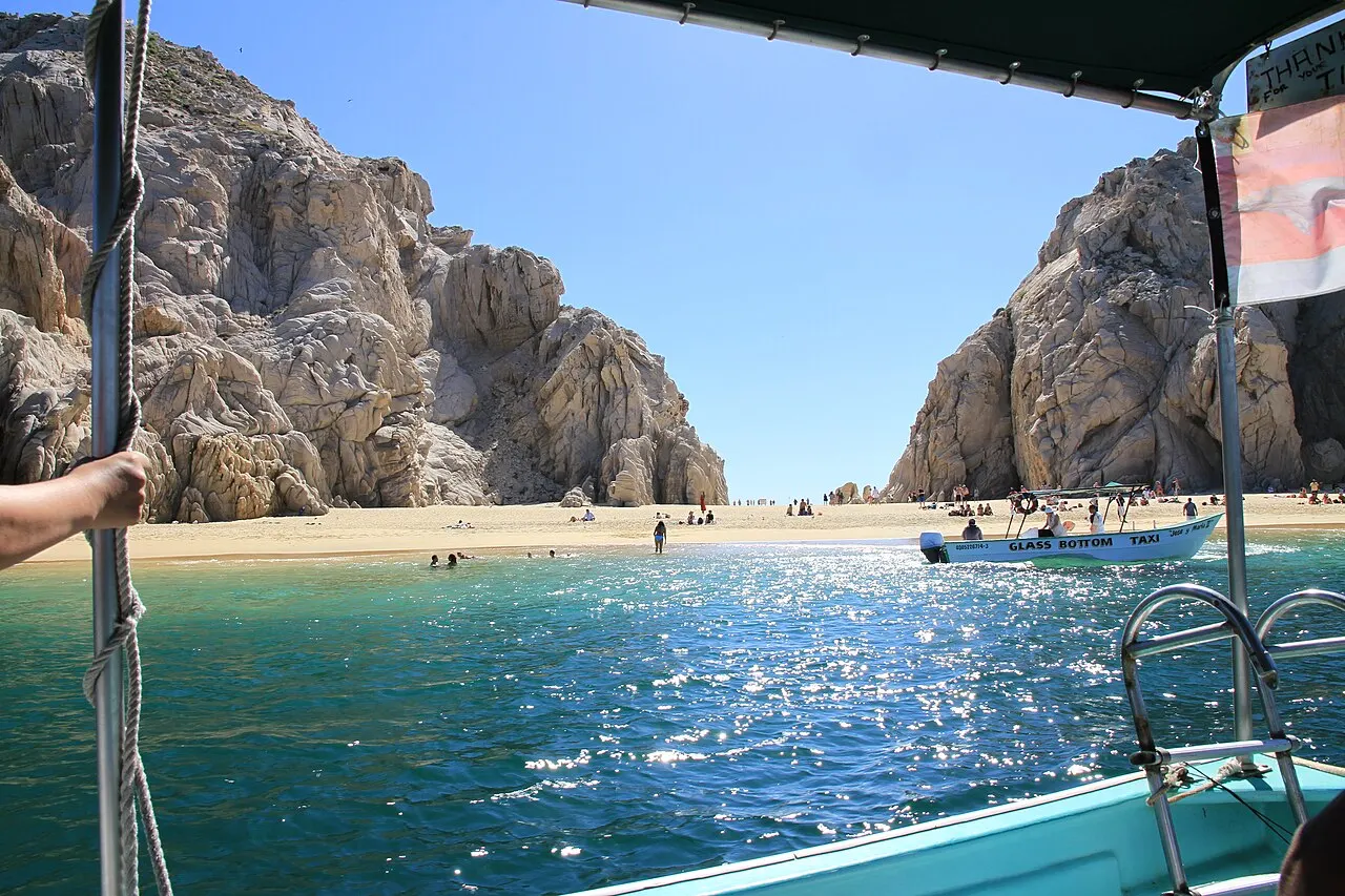 Faixa de areia entre dois paredões de rocha, com água azul brilhando e um barco ancorado, vista da proa de outro barco.