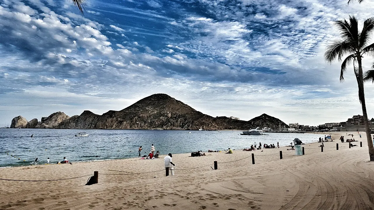 Praia larga de areia com poucas pessoas, a baía calma e morros rochosos no horizonte, sob céu com nuvens e uma palmeira à direita.