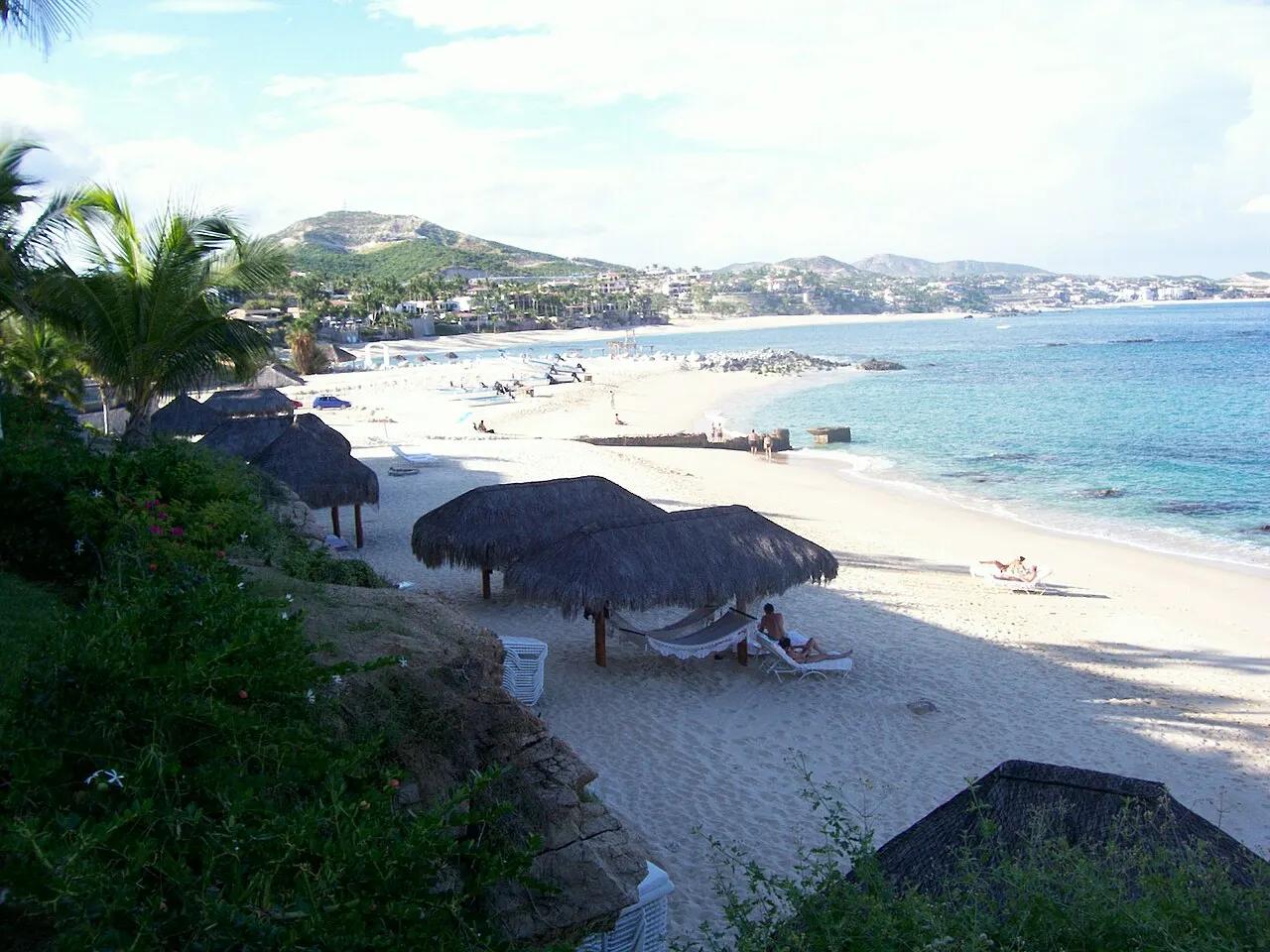 Praia de areia branca com guarda-sóis de palha e espreguiçadeiras, vista do alto entre palmeiras, com mar calmo e morros com casas ao fundo.
