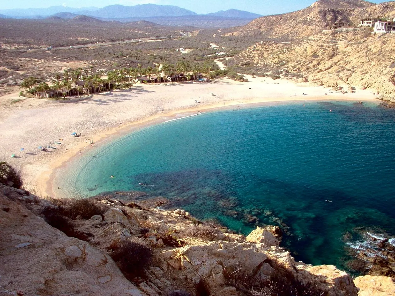 Enseada em meia-lua de água turquesa e areia clara, vista do alto de rochas, com morros secos e serras ao fundo.