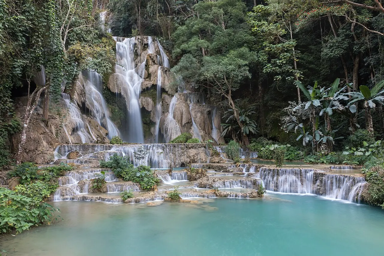 A queda principal de Kuang Si descendo pelo calcário entre a mata, e na frente degraus de pequenas cascatas que terminam numa piscina de água turquesa.