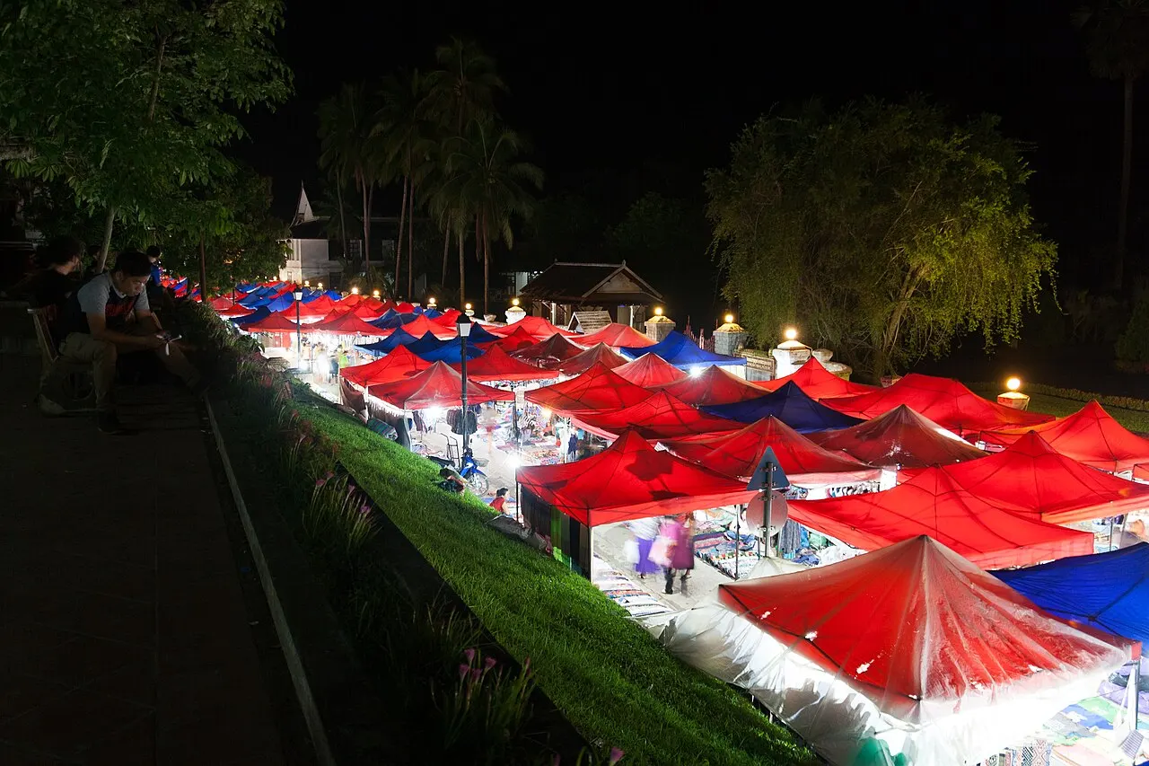 Rows of stalls under red and blue awnings, lit from inside, along a street at night, seen from a higher lawn with dark trees behind.
