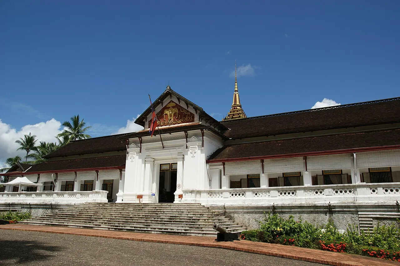 The long white Royal Palace building, with a dark roof and an entrance under a gilded gable, at the top of a wide staircase, with a golden temple spire behind and a blue sky.