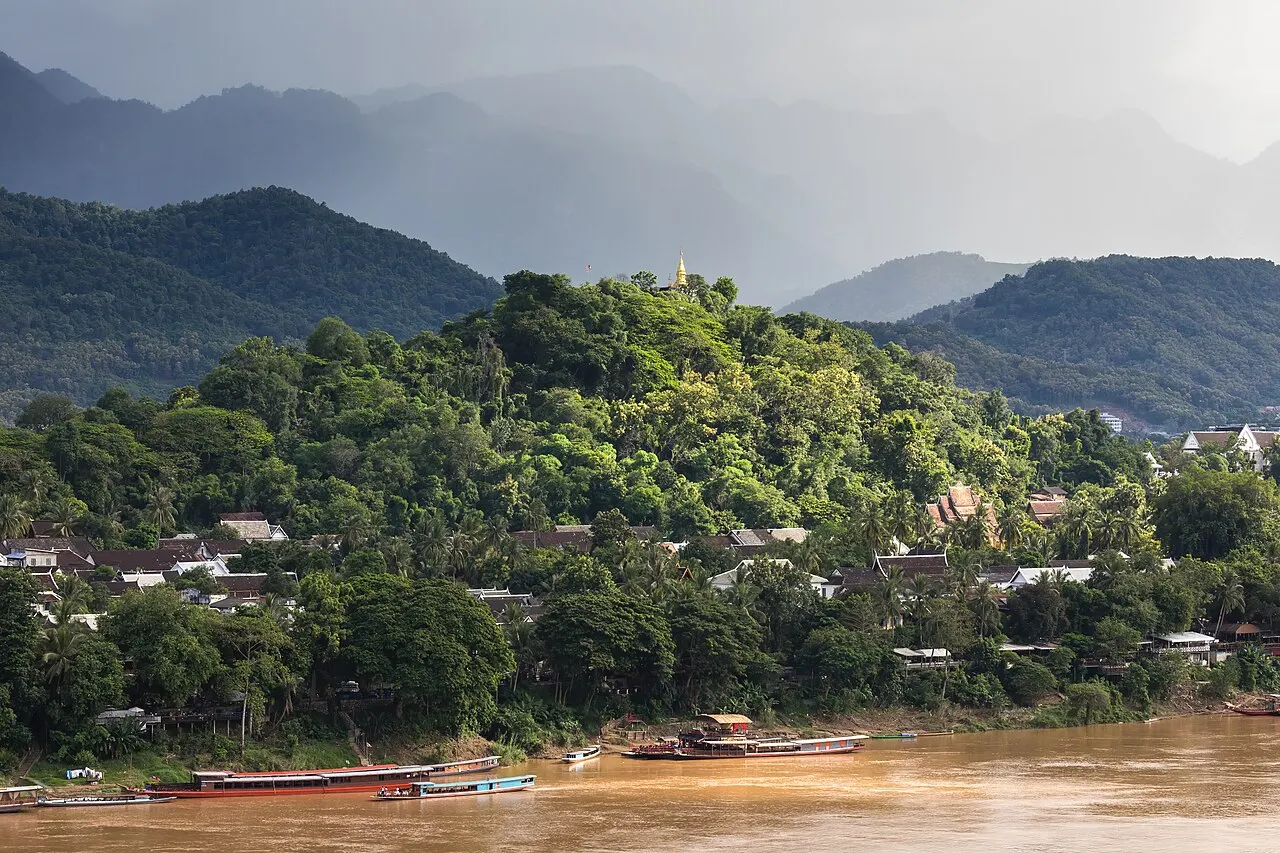 O morro Phou Si, coberto de árvores e com um pequeno stupa dourado no topo, sobre as casas da cidade, visto do outro lado do rio Mekong, com barcos compridos na margem e montanhas ao fundo.