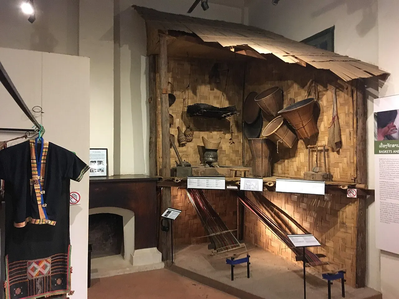 Uma sala do museu com a reconstrução de uma parede de casa de bambu trançado, com cestos e ferramentas pendurados, teares de madeira no chão e uma roupa escura bordada exposta à esquerda.