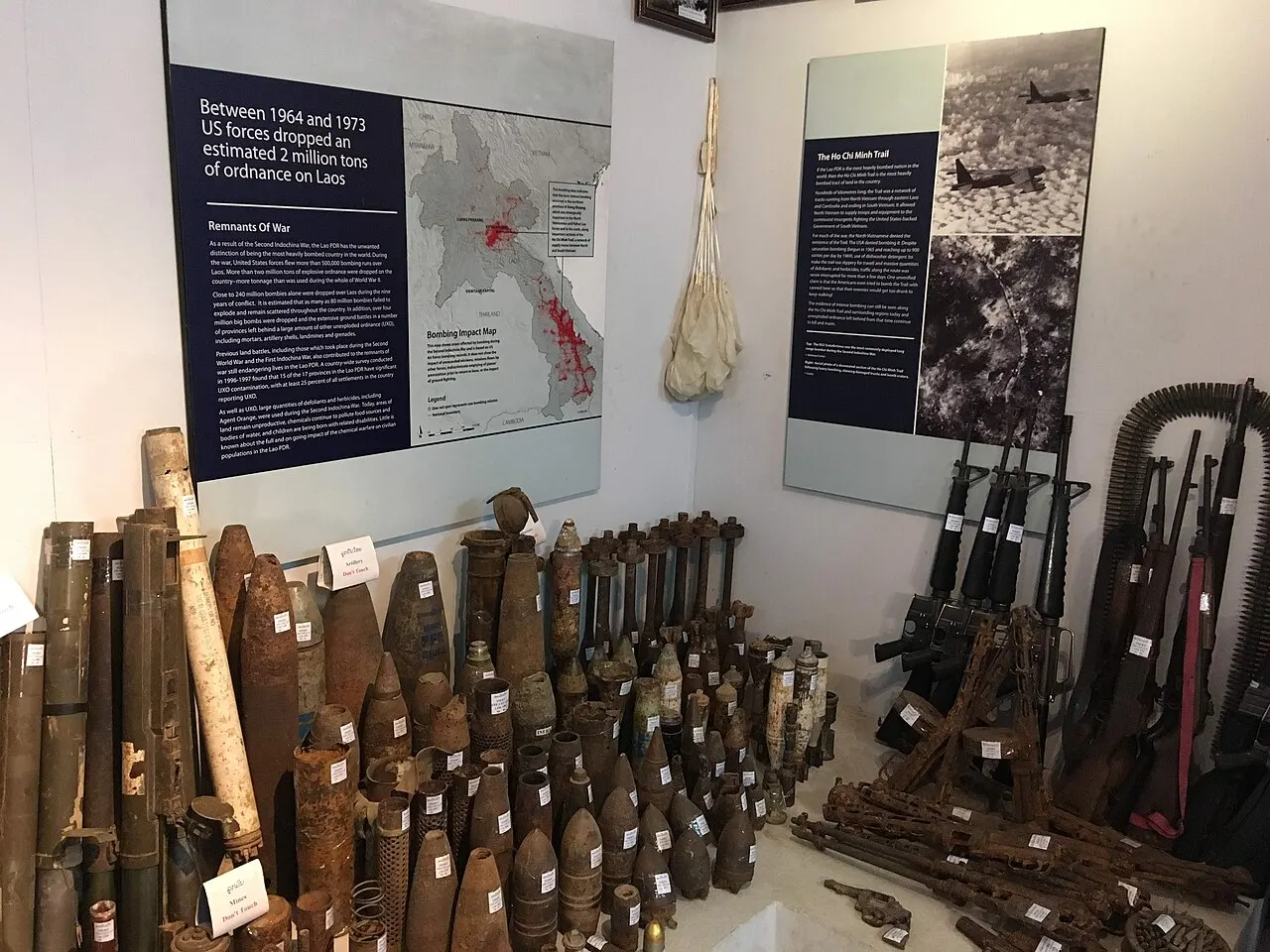 Um canto da sala de exposição com dezenas de invólucros de bombas e munições enferrujados no chão, armas encostadas na parede e painéis com um mapa do Laos e fotografias aéreas.