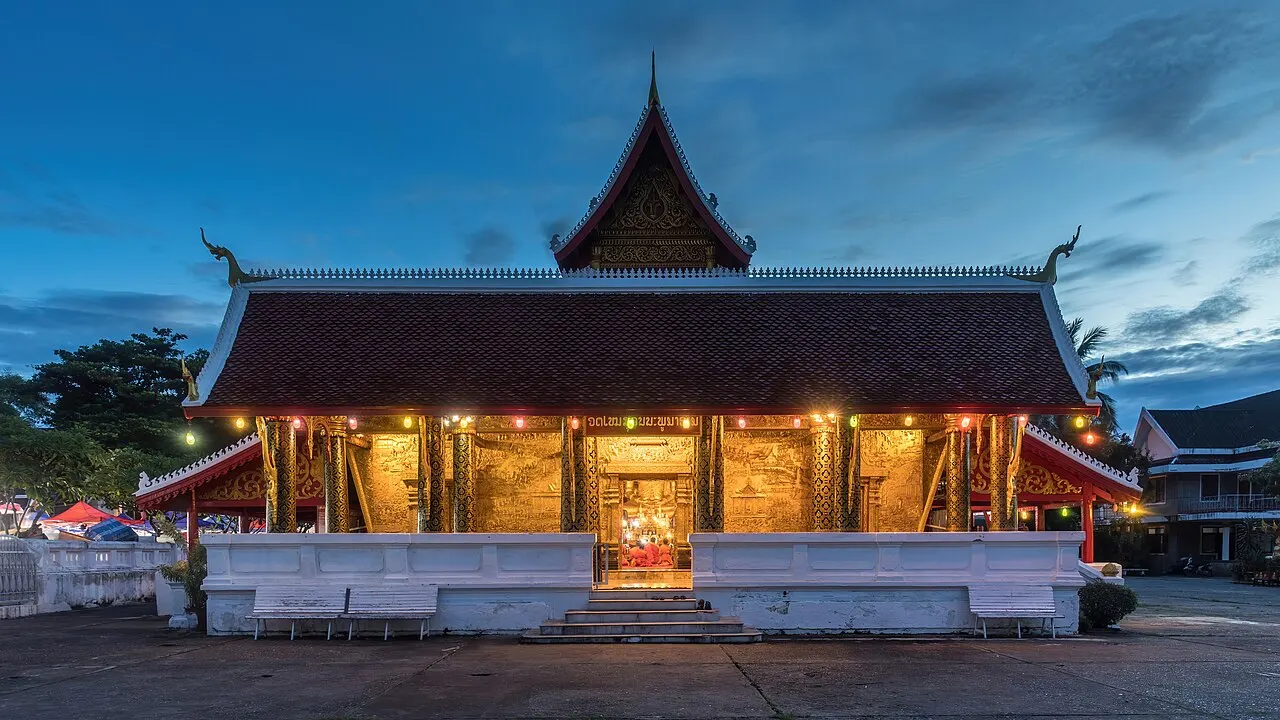 The front of Wat Mai at dusk, with its layered roof and a gilded porch lit from inside, on a white base under a deep blue sky.