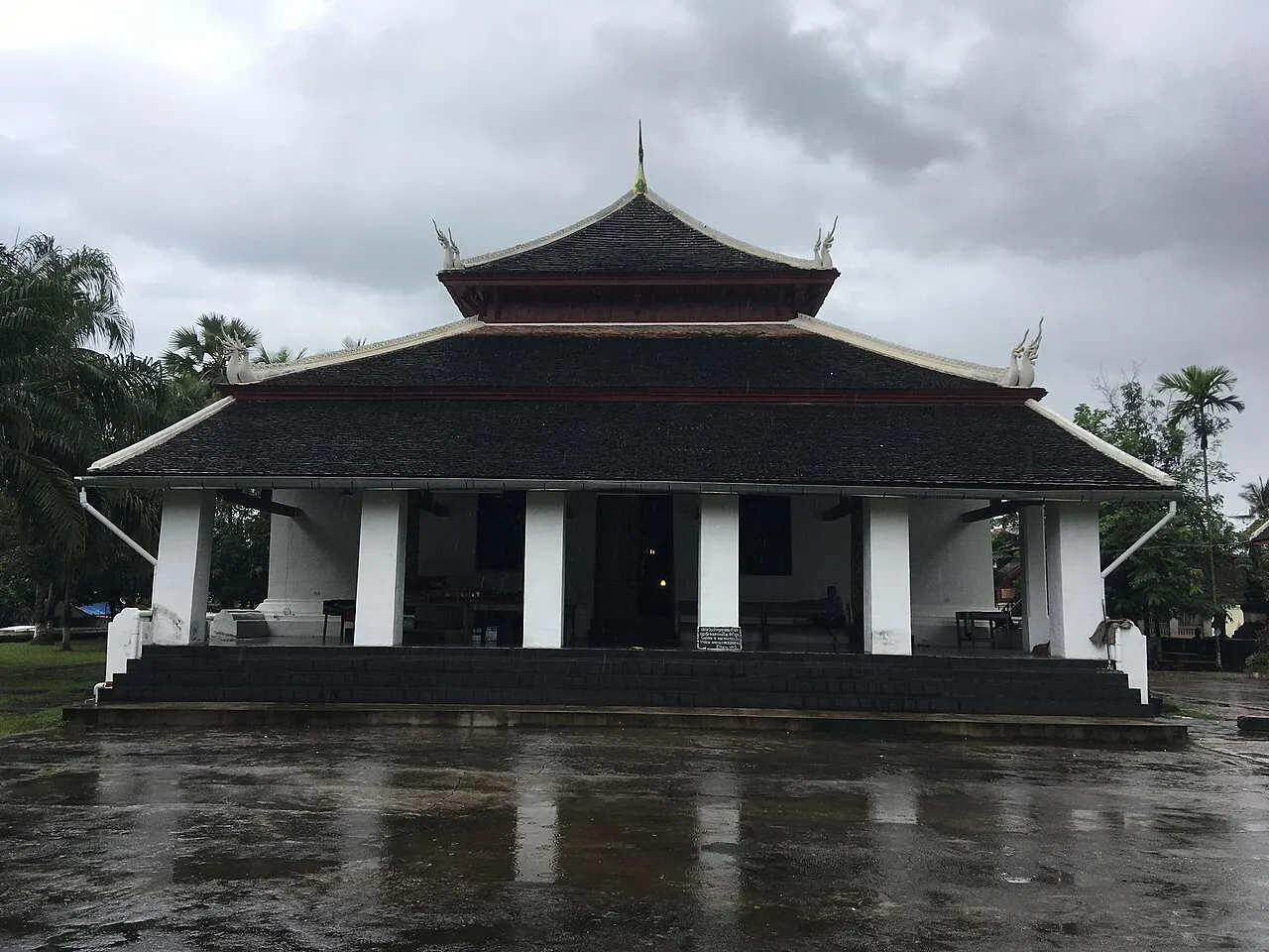 The front of Wat Visounnarath, with a dark two-tier roof and a porch of white columns, seen head-on across a rain-wet courtyard under a cloudy sky.