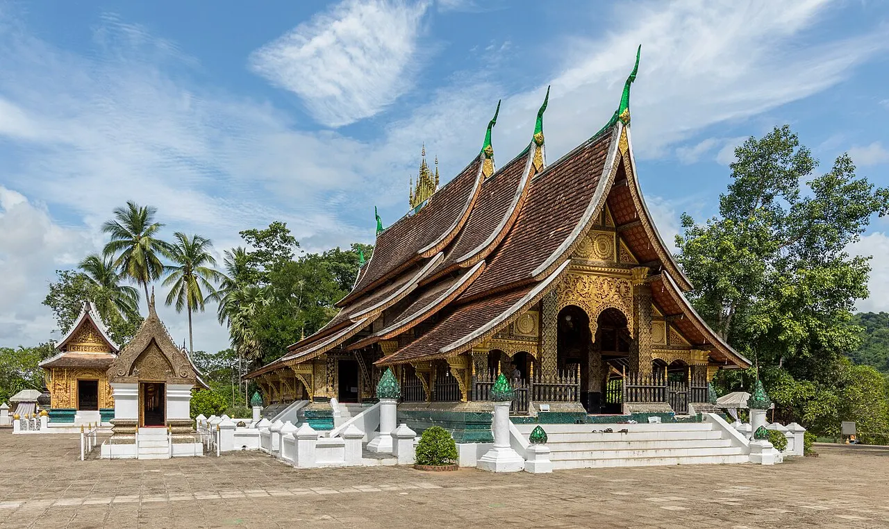 O templo de Wat Xieng Thong, com telhados escuros em camadas que descem quase até o chão e frontão dourado, ao lado de duas capelas menores, sob céu azul com palmeiras.