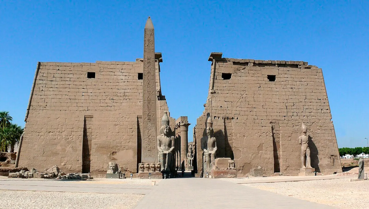 Pilone de entrada do Templo de Luxor, com o obelisco de granito e os colossos sentados, sob céu azul.
