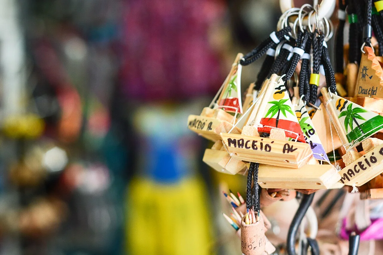 Chaveiros de madeira em forma de jangada, pintados com coqueiros e a inscrição Maceió AL, pendurados numa banca de artesanato com o fundo desfocado.