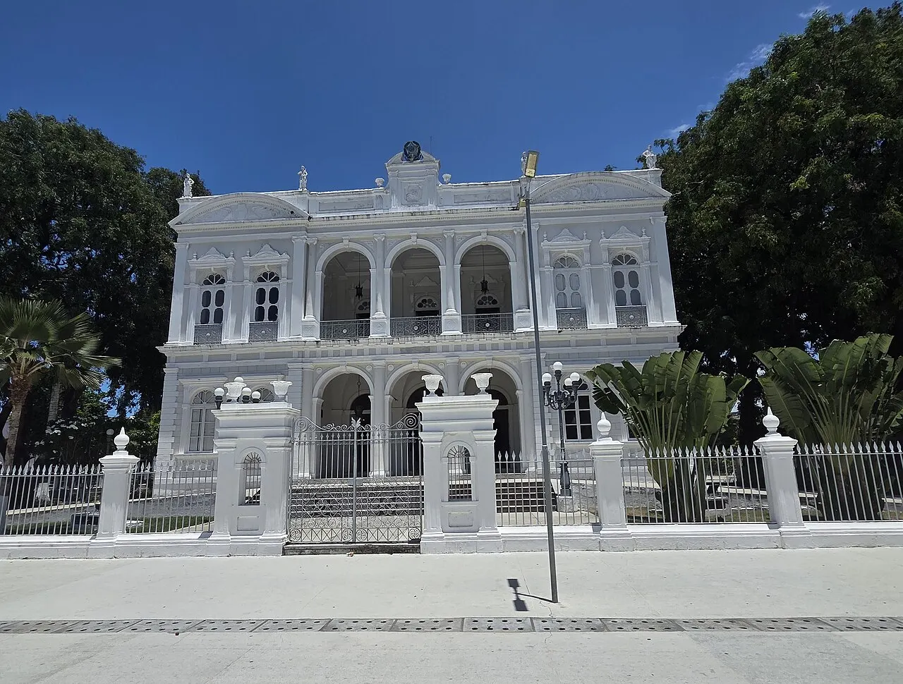 A fachada branca e simétrica do Palácio Floriano Peixoto, com dois andares de arcos e o portão de grade, entre árvores, sob céu azul.