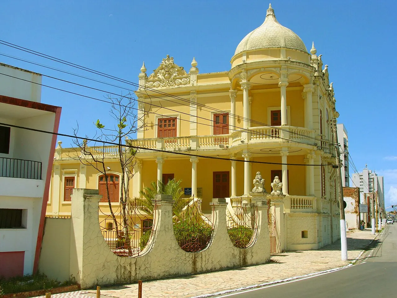O palacete amarelo do Museu Théo Brandão, com cúpula, varandas em arco e muro baixo de balaústres, numa esquina sob céu azul.