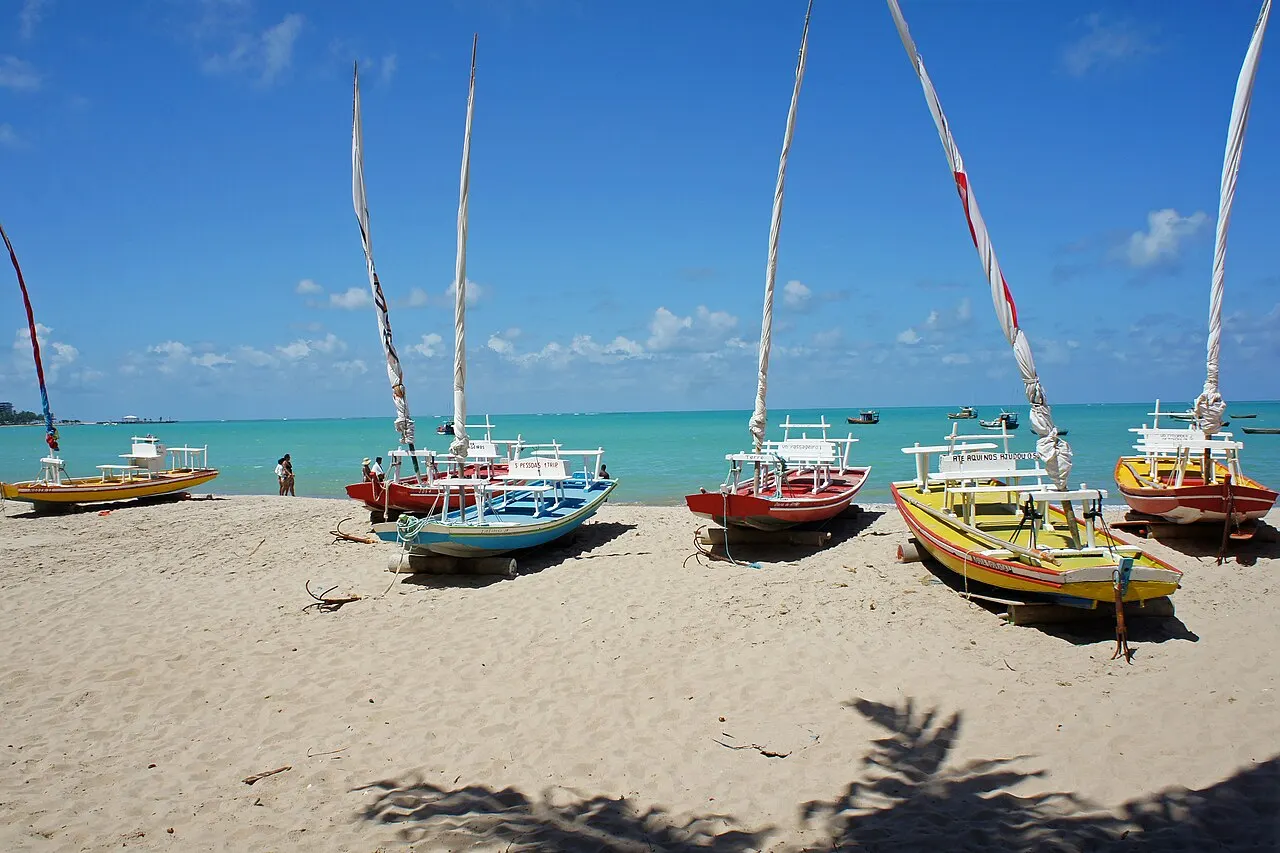 Jangadas coloridas de vela alta paradas na areia da praia da Pajuçara, com o mar verde e raso atrás e céu azul.
