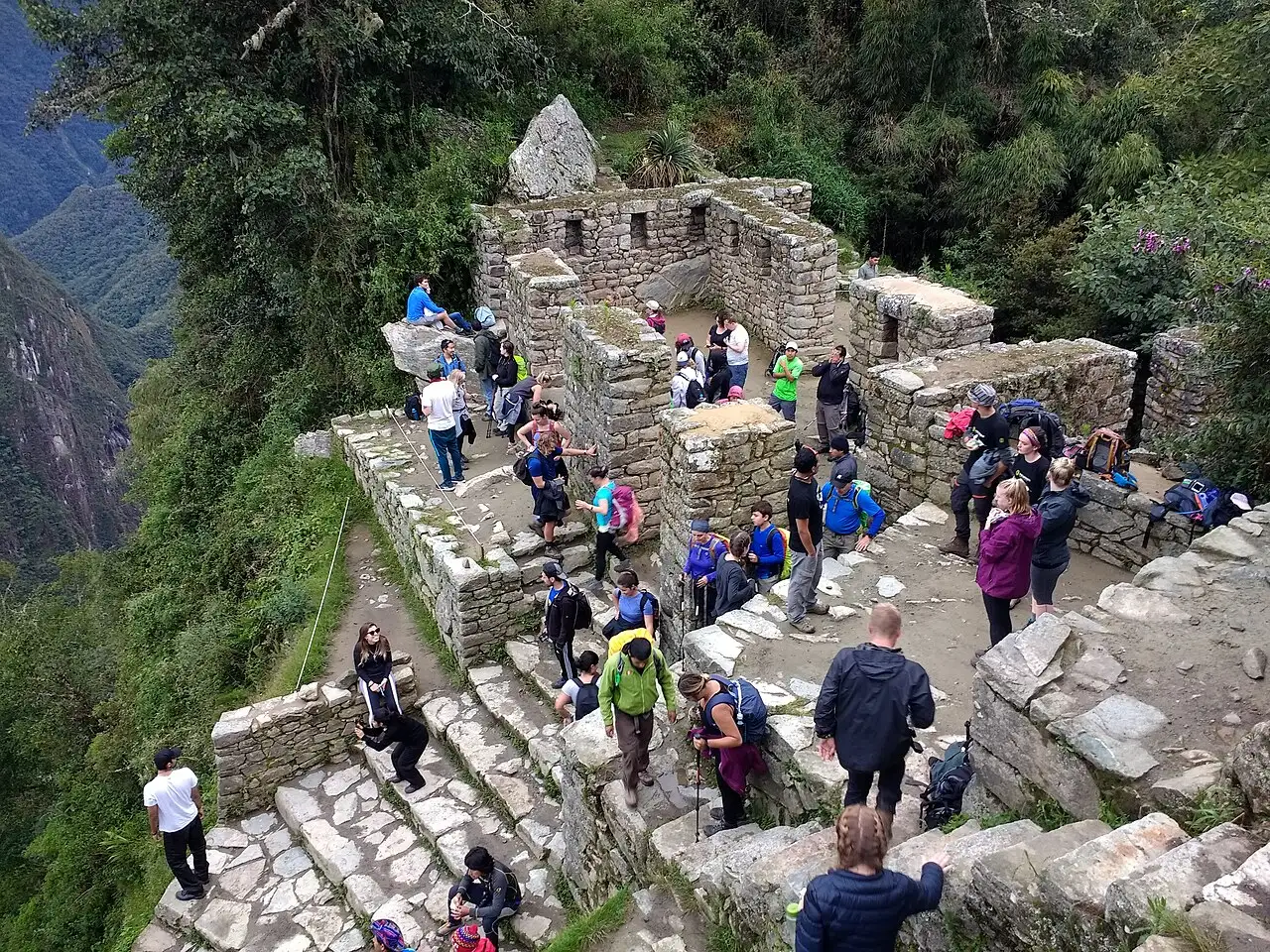 Intipunku na crista, com as paredes de pedra, os degraus e pessoas de mochila, e o vale ao lado.