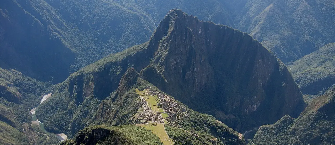 A cidadela de Machu Picchu vista do alto, com terraços verdes, o Huayna Picchu e encostas arborizadas ao fundo.